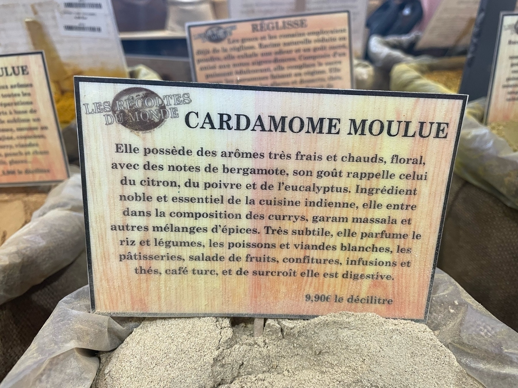 Label describing ground cardamom, with aromatic profile and culinary uses, set against a backdrop of spices.
Text transcription: LES ÉPICES DU MONDE CARDAMOME MOULUE
Elle possède des arômes très frais et chauds, floral, avec des notes de bergamote, son goût rappelle celui du citron, du poivre et de l'eucalyptus. Ingrédient noble et essentiel de la cuisine indienne, elle entre dans la composition des currys, garam massala et autres mélanges d’épices. Très subtile, elle parfume le riz et légumes, les poissons et viandes blanches, les pâtisseries, salade de fruits, confitures, infusions et thés, café turc, et de surcroît elle est digestive. 9,90€ le décilitre