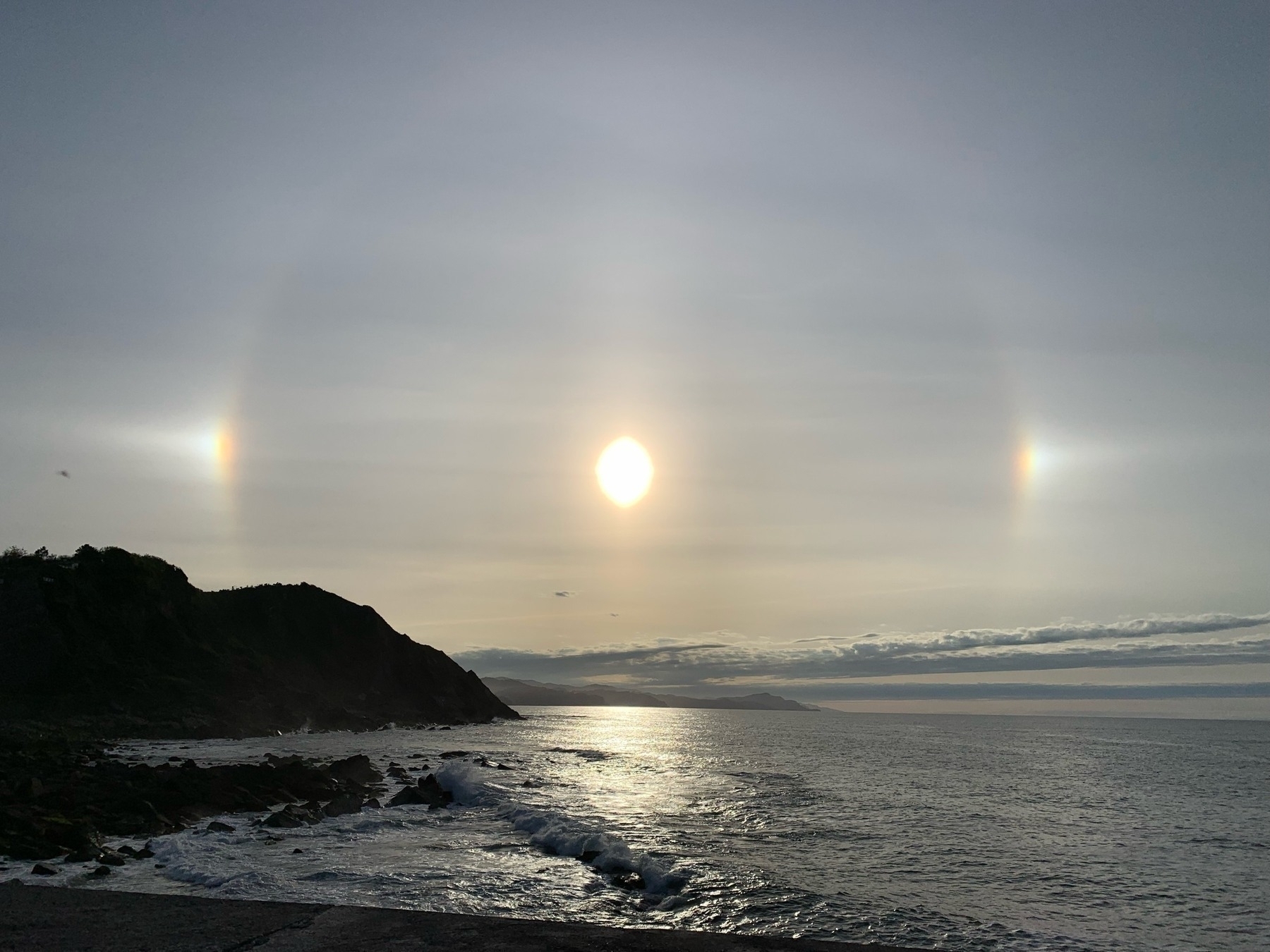 Sun with halo phenomenon over the sea, flanked by two faint sun dogs, at coastline during twilight.
Spanish: Sol con fenómeno de halo sobre el mar, flanqueado por dos parhelios tenues, en la costa durante el crepúsculo.
Basque: Eguzkia halo fenomenoarekin itsasoaren gainean, bi izpi ahul albo batean, kostaldean ilunabarrean.