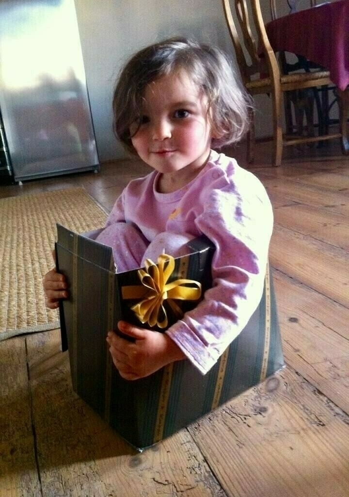 A young child sits inside a large gift box, looking at the camera, in a homely room with wooden flooring.
Spanish: Un niño pequeño se sienta dentro de una caja de regalo grande, mirando a la cámara, en una habitación acogedora con suelo de madera.
Basque: Haur txiki bat opari-kutxa handi baten barruan eserita dago, kamera begiratzen, egurrezko zorua duen etxe-giroko gelan.