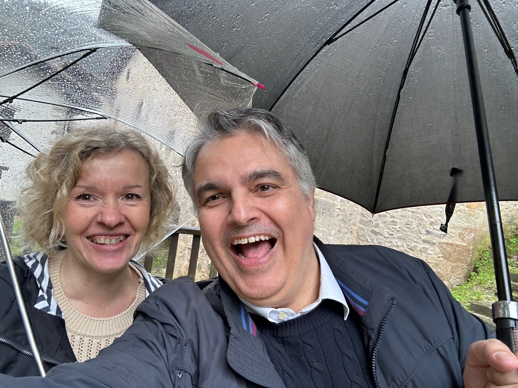 Two people smiling under umbrellas in a rainy setting with a building visible in the background.