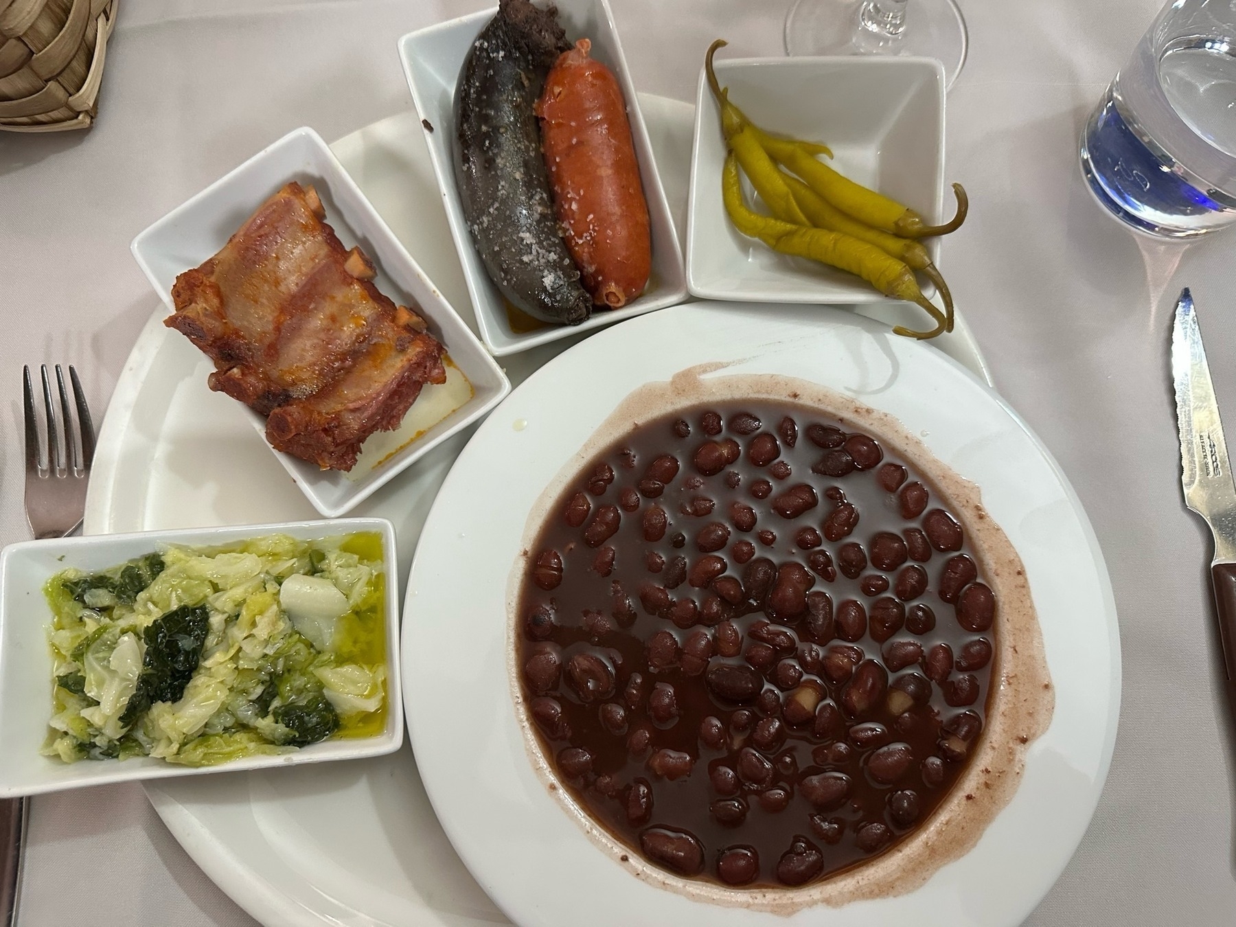 A plate of dark beans in broth is surrounded by side dishes of cabbage salted meats and green peppers on a white tablecloth next to a fork and knife.