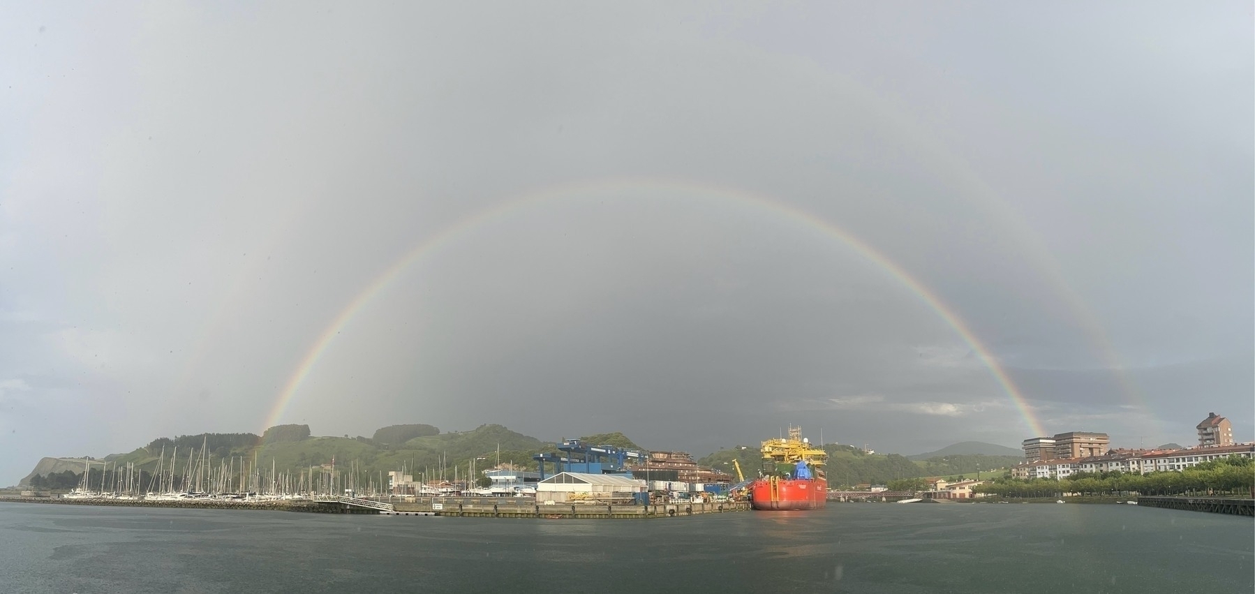 A double rainbow arches over a harbor with moored sailboats, industrial equipment, and distant hills.
Spanish: Un doble arcoíris se extiende sobre un puerto con veleros amarrados, equipos industriales y colinas lejanas.
Basque: Halabehar bikoitz bat portu batean zehar makurtzen da, itsasontzi bela itsasontziak, ekipamendu industrialak eta urrutiko muinoak dituela.