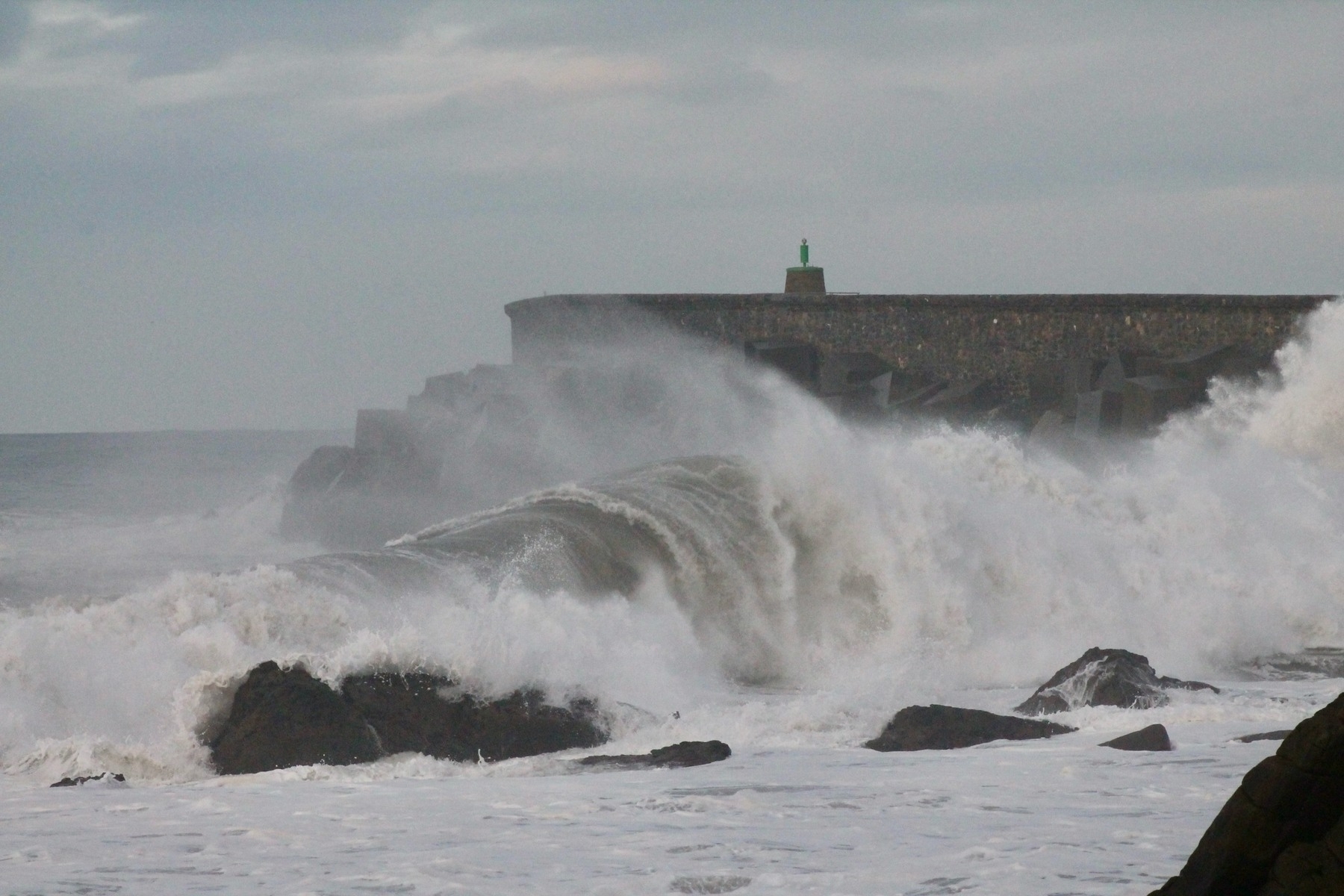 Large waves crash against rocks near a seawall, under a cloudy sky.
Spanish: Grandes olas chocan contra las rocas cerca de un malecón, bajo un cielo nublado.
Basque: Olatu handiak harrien aurka kraskatzen ari dira, malekon baten ondoan, zeru hodeitsu baten azpian.