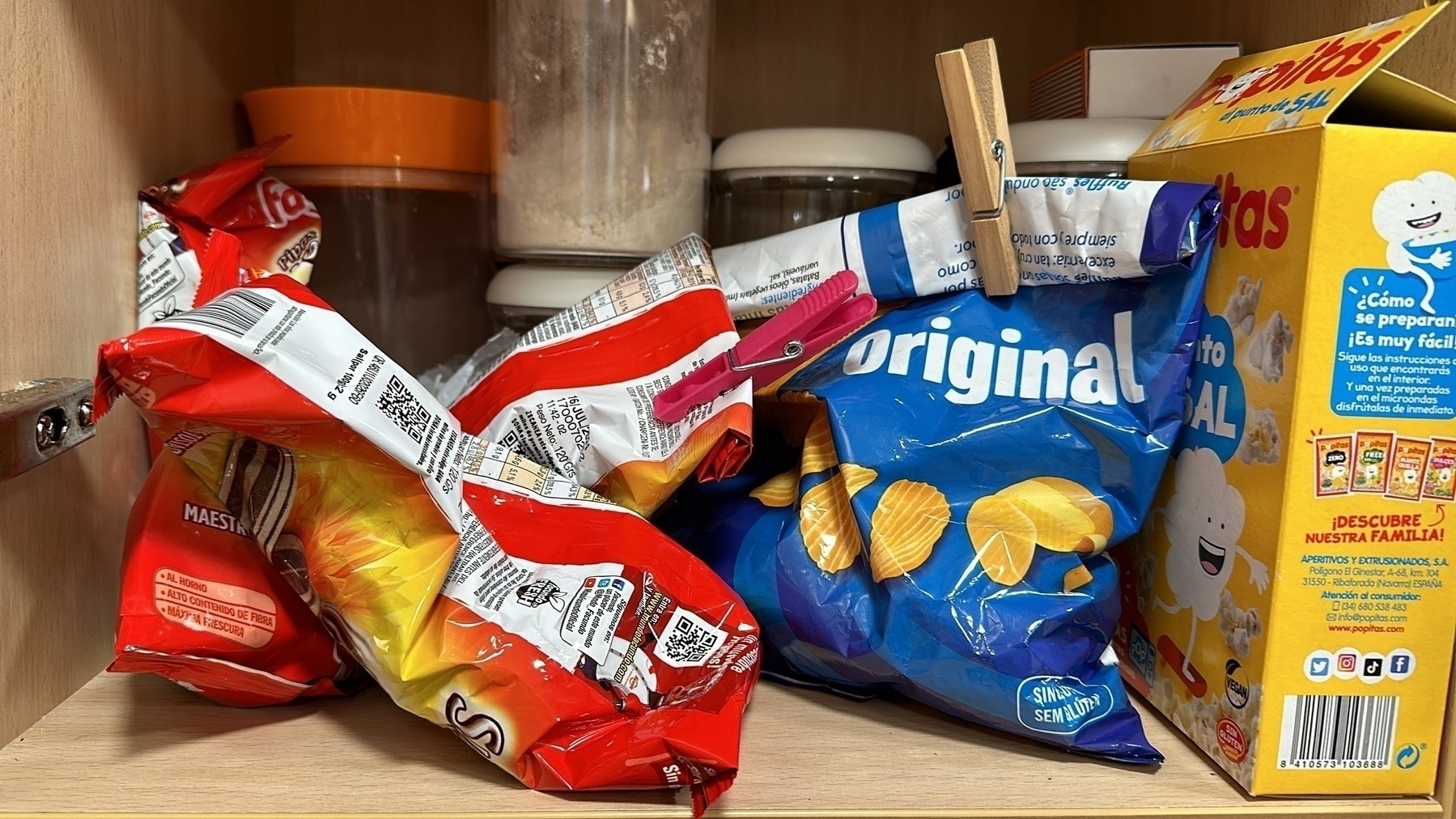 Several snack bags and a cereal box are cluttered on a wooden pantry shelf among kitchen items.
Spanish: Varias bolsas de snacks y una caja de cereales están amontonadas en un estante de despensa de madera entre artículos de cocina.
Basque: Hainbat snack poltsa eta zereal kutxa bat trasteleku batean daude bilduta sukaldeko tresnen artean, egurrezko panategi-apal batean.