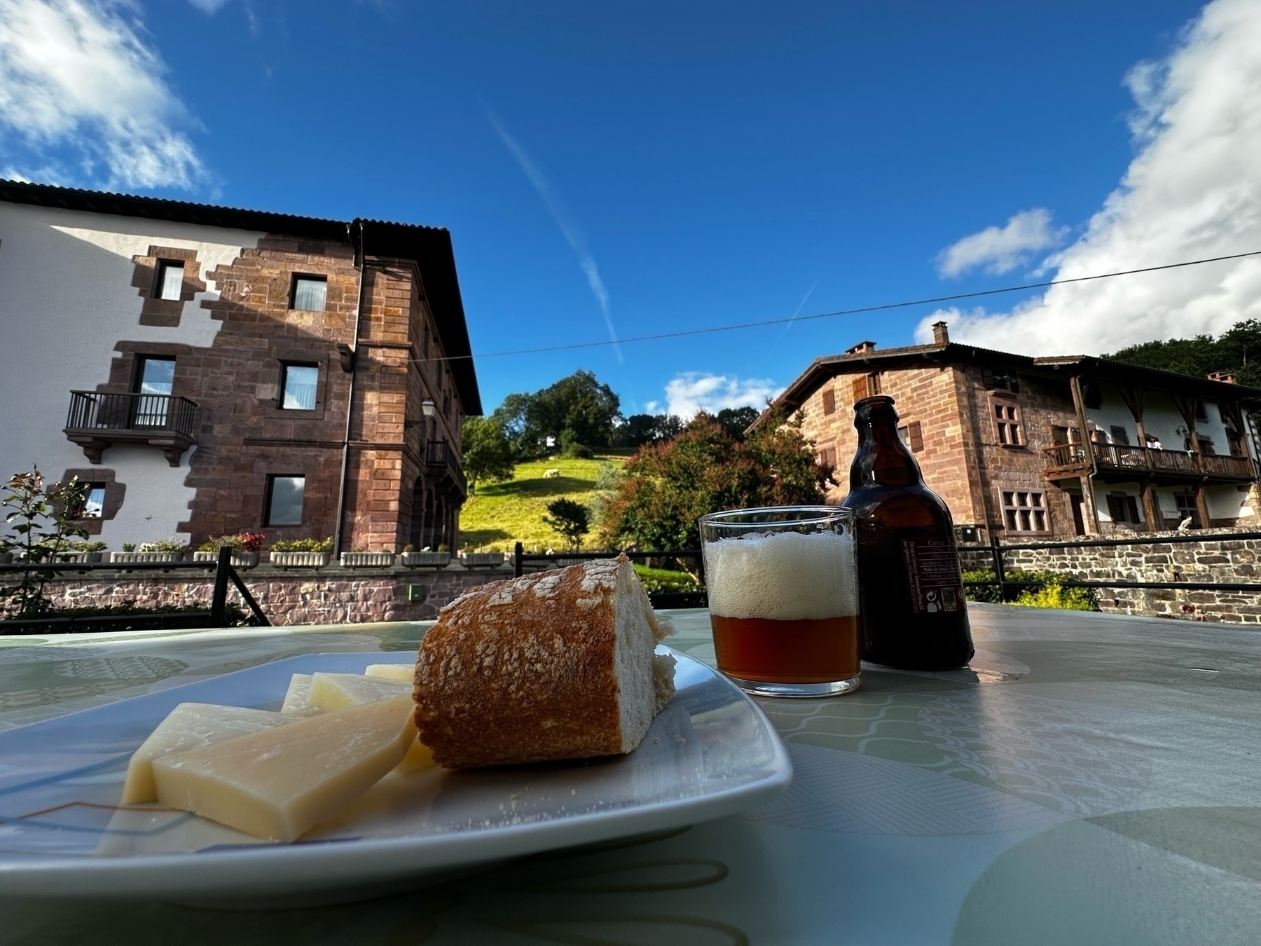 A plate of cheese and bread with a beer bottle and glass sits on an outdoor table, traditional stone houses against a backdrop of green hills and a blue sky.
Spanish: Un plato de queso y pan con una botella de cerveza y vaso están sobre una mesa al aire libre, casas de piedra tradicionales contra un telón de fondo de colinas verdes y cielo azul.
Basque: Gazta eta ogi plater bat garagardo botila eta begikoa mahai baten gainean dago, atzean etxe harritsu tradizionalak, berde koloreko muinoak eta zeru urdin bat dituela.