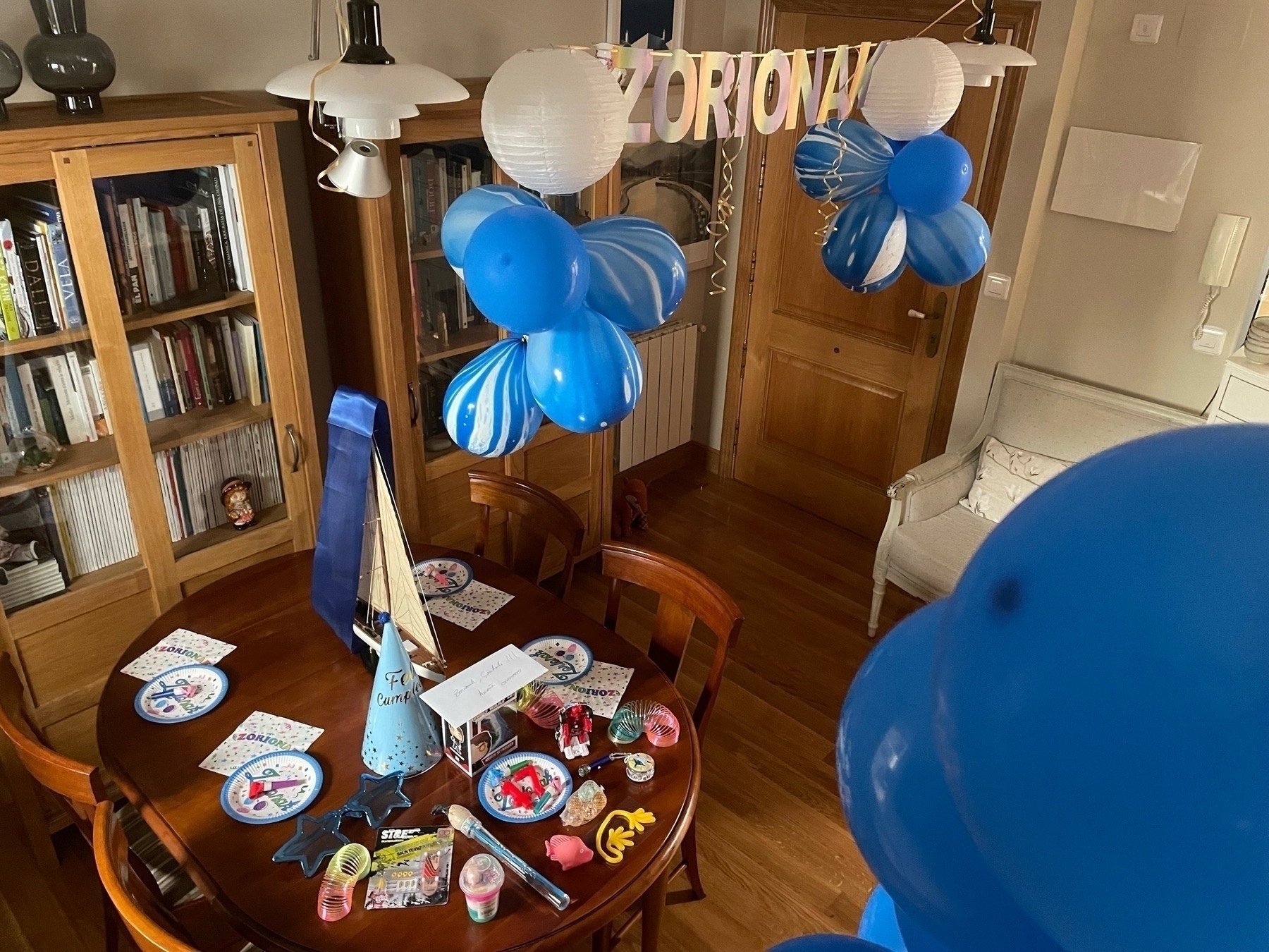 A birthday table is set with decorations, favors, and blue balloons in a home interior; a banner reads ZORIONAK, congratulations in Basque.