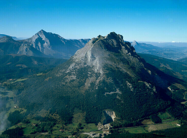 A jagged mountain peak rises above a forested valley under a clear blue sky.
Spanish: Un pico montañoso dentado se eleva sobre un valle boscoso bajo un cielo azul claro.
Basque: Harri-zorrotz mendi-gailur bat ageri da baso estalitako haranaren gainetik zeru urdin argi baten azpian.