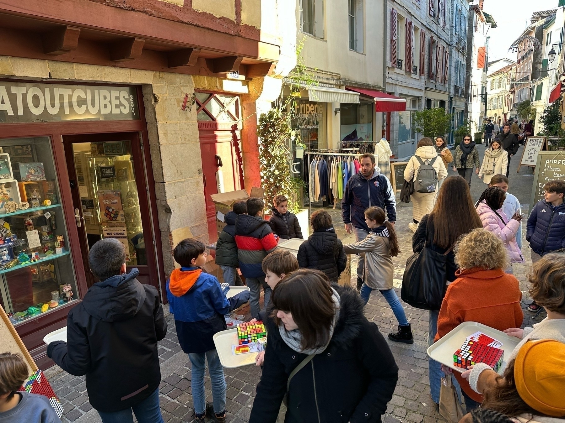 Children gather in a cobblestone street, some playing with Rubik's Cubes, outside a puzzle shop in a quaint town setting.
Spanish: Niños y niñas reunidas en una calle empedrada, algunas jugando con Cubos de Rubik, frente a una tienda de rompecabezas en un entorno pintoresco de pueblo.
Basque: Haurrak bildu dira harrizko kale batean, batzuk Rubik kuboekin jolasten, herriko inguru xarmagarri batean dagoen puzzle denda baten kanpoaldean.