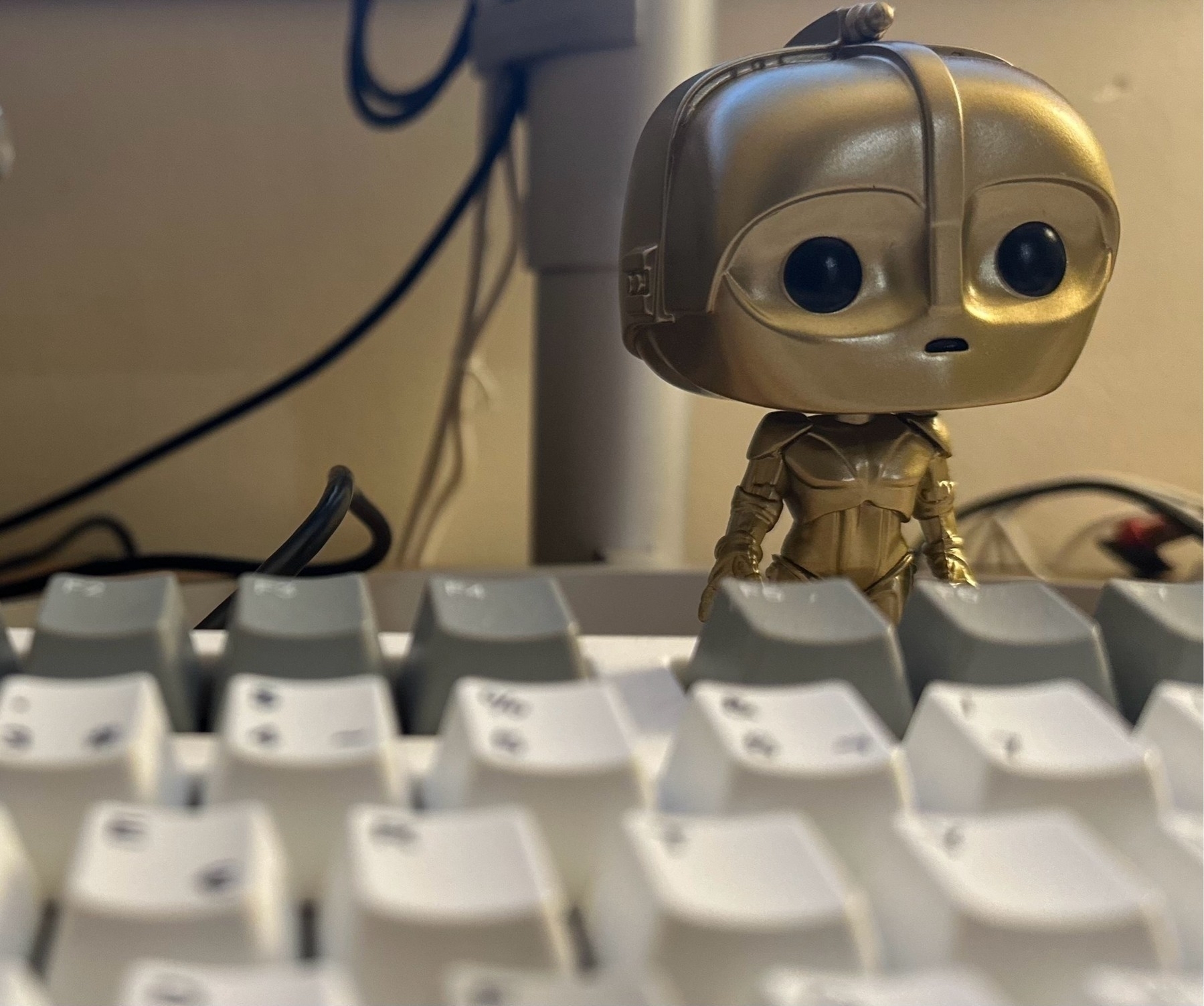 A C-3PO funko figurine with large eyes stands on a keyboard; cables and a monitor in the background.
