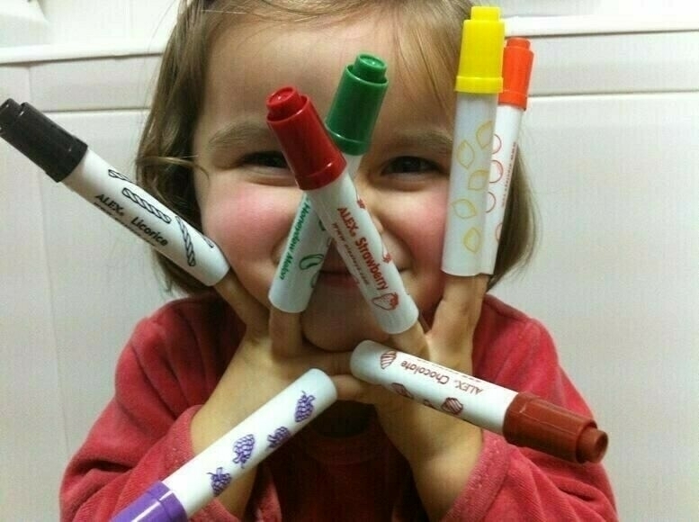 A smiling child holds up multiple colorful markers playfully arranged around their face, in a white-tiled room.
Spanish: Un niño sonriente sostiene múltiples marcadores de colores, dispuestos de forma juguetona alrededor de su rostro, en una habitación con azulejos blancos.
Basque: Aurpegian koloretsuak diren hainbat markatzaile jolasean antolatuta dituen haur irribarretsu bat, zurezko gelan.